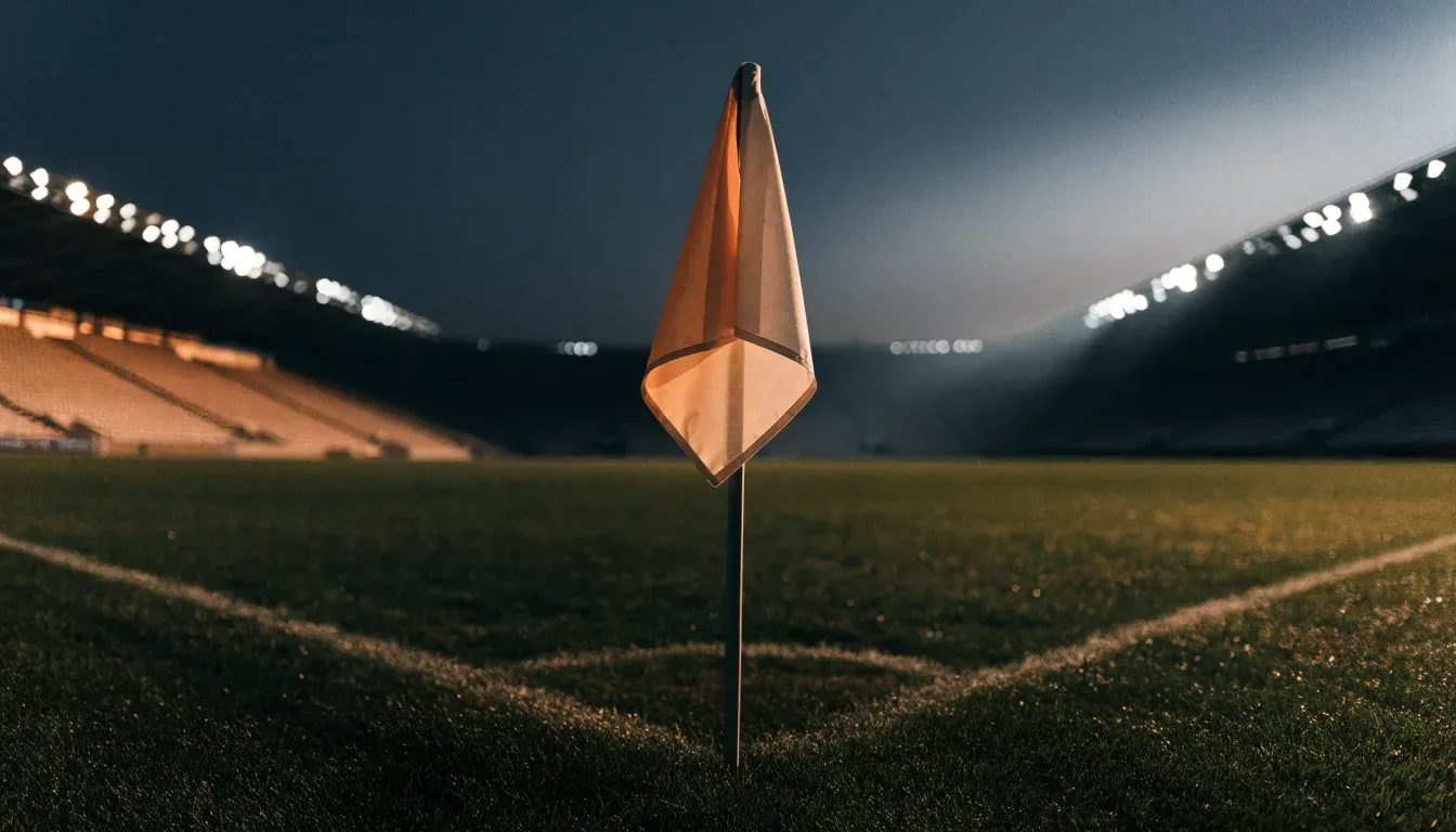 Eine Eckfahne am Rand eines Rasenplatzes, dahinter ein leeres Stadion unter Flutlicht aus der Ferne gesehen.