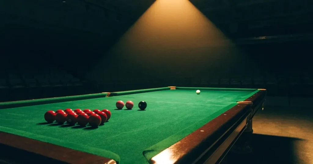 Ein Snookertisch mit grünem Tuch in einem abgedunkelten Saal, ein Lichtkegel fällt von oben auf die aufgestellten Kugeln.