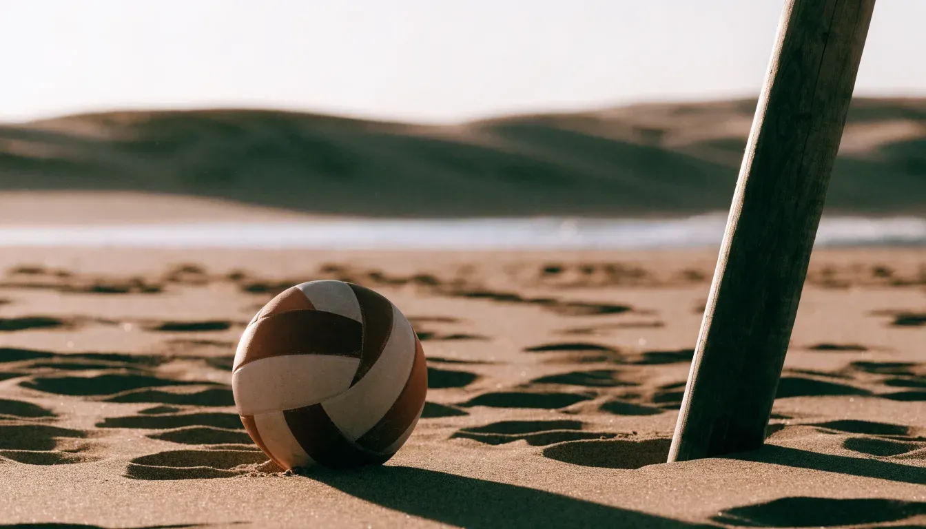 Ein Volleyball liegt auf Sand neben einem Netzpfosten, Strandlicht am späten Nachmittag, im Hintergrund unscharfe Dünen.